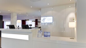 Un espace d’accueil de l’hôtel Holiday Inn Express, doté d’un design moderne, comprenant un comptoir d’accueil, un écran numérique affichant le nom de l’hôtel et un panneau indiquant le programme de fidélité Priority Club.