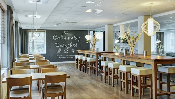 A restaurant with a long wooden table and chairs, a blackboard with 'Culinary Delights' written on it, and a view of the city through large windows. - Holiday Inn Express Amsterdam - Schiphol by IHG (Hoofddorp)