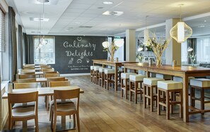 Un restaurant avec une longue table et des chaises en bois, un tableau noir sur lequel est écrit « Délices culinaires », et une vue sur la ville grâce à de grandes fenêtres.