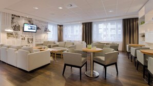 Un hall d’hôtel moderne, doté d’un espace salon, d’un téléviseur fixé au mur et de grandes fenêtres avec des rideaux.