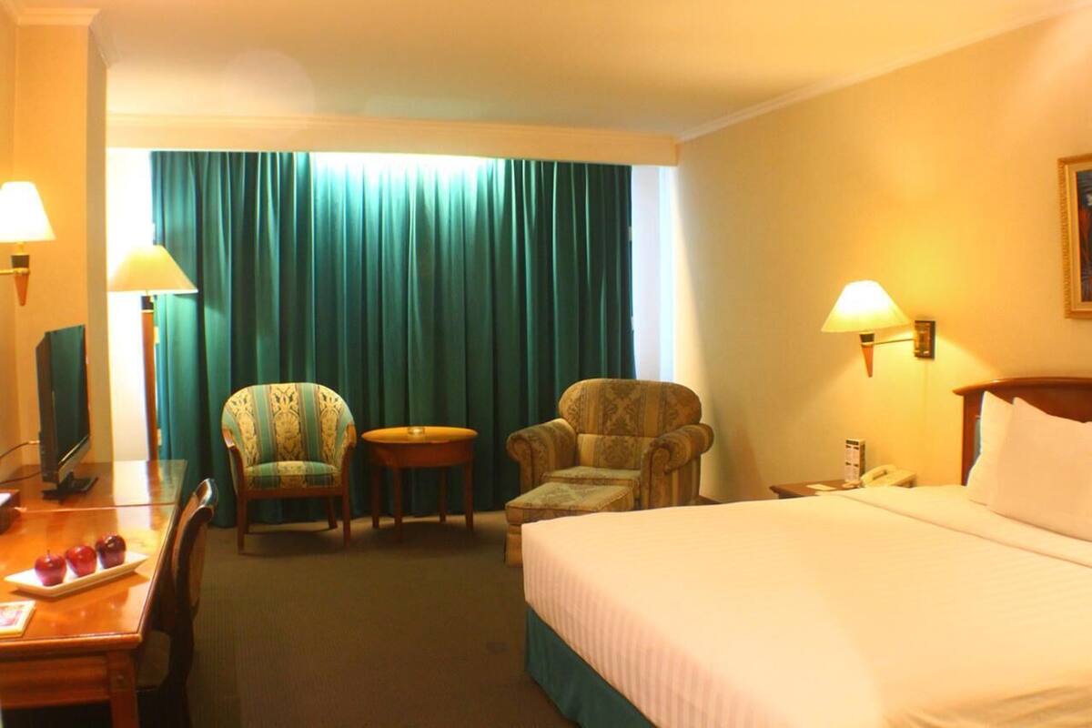 Hotellihuone, jossa on sänky, kaksi nojatuolia, pieni pöytä, työpöytä lampun kanssa ja televisio.