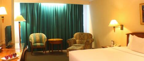 A hotel room with a bed, two armchairs, a small table, a desk with a lamp, and a television.