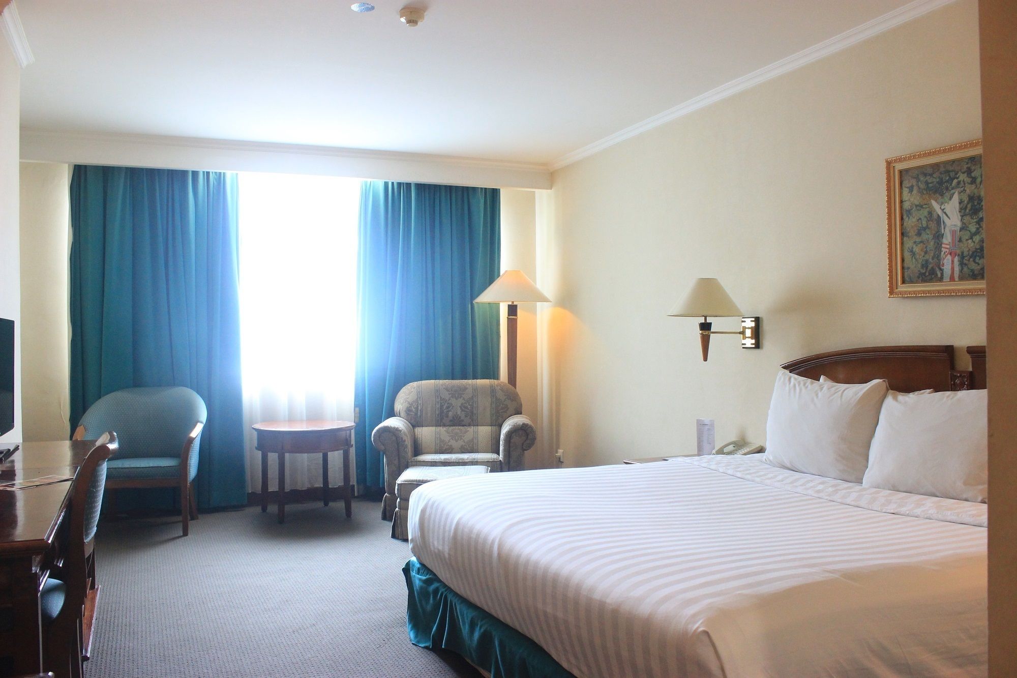 A hotel room with a bed, two armchairs, a small table, a desk, a lamp, and a painting on the wall.