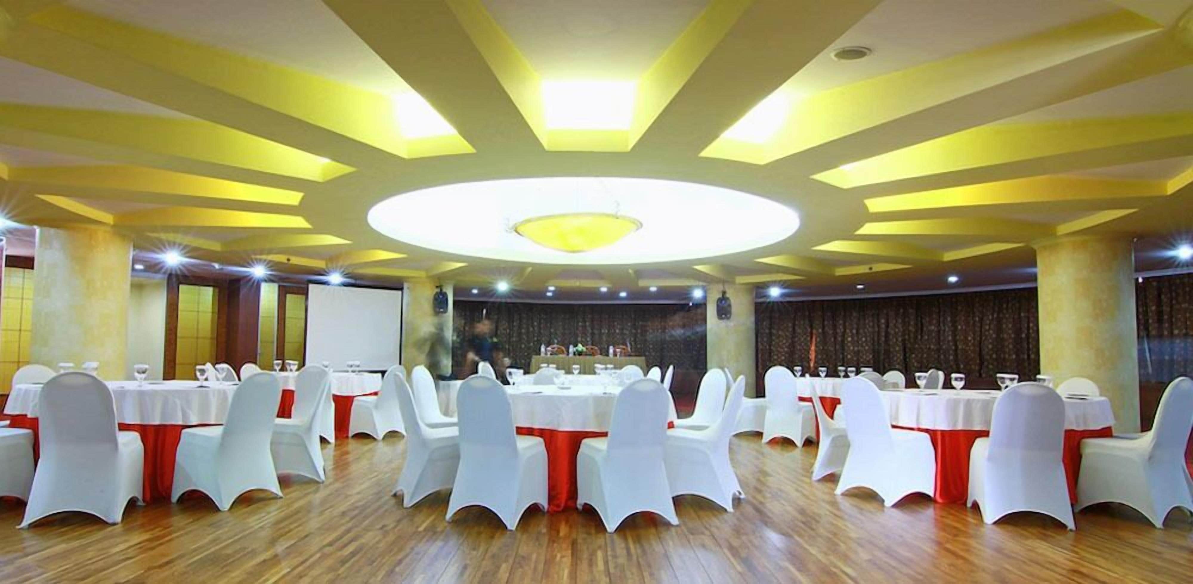 Een feestzaal met ronde tafels, witte stoelen en een centrale ronde plafondlamp.