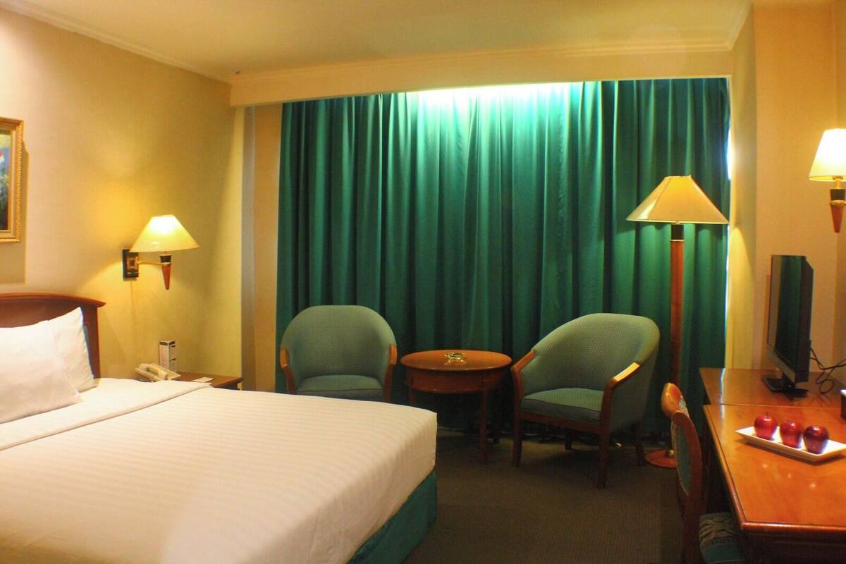 Hotellihuone, jossa on sänky, kaksi nojatuolia, pieni pöytä, työpöytä tietokoneella ja televisio.