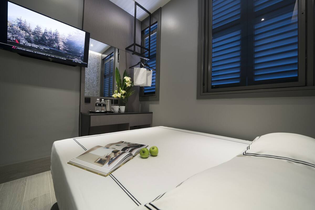 Een moderne hotelkamer met een groot bed, een flatscreen televisie aan de muur, een klein tafeltje met een bloevas en een raam met luxaflex.