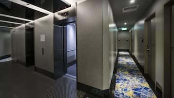A hotel corridor with a patterned carpet, a door with a digital display, and a wall-mounted panel.
