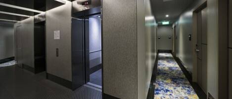 A hotel corridor with a patterned carpet, a door with a digital display, and a wall-mounted panel.