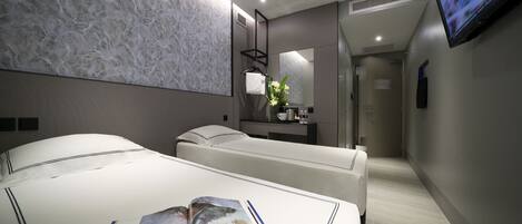 Una habitación de hotel moderna con una cama grande, una mesita de noche con una revista y manzanas, un pequeño rincón de descanso, un televisor y un baño con espejo y lavamanos.
