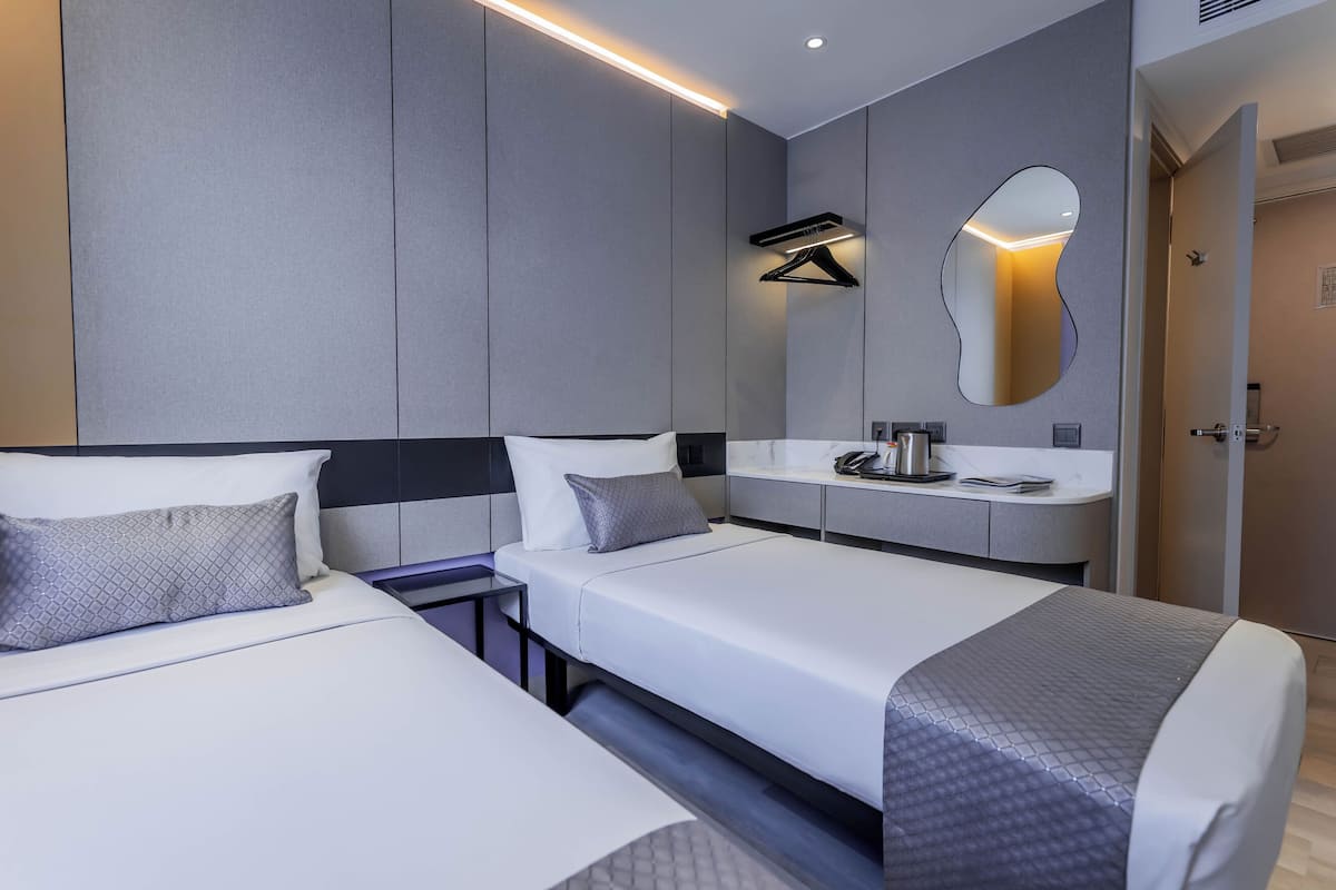 Quarto de hotel moderno com duas camas de solteiro, mesa de cabeceira com abajur, espelho grande e toucador embutido com pia e espelho.