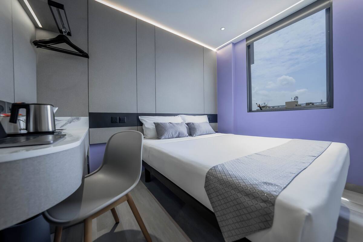 Quarto de hotel moderno com uma cama, uma mesa com chaleira, uma cadeira e uma janela grande com vista para a cidade.
