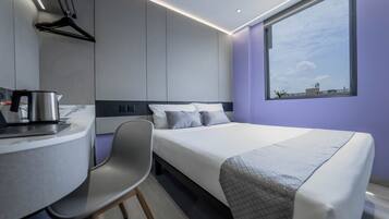 Una habitación de hotel moderna con una cama, un escritorio con hervidor, una silla y un ventanal que ofrece vistas a la ciudad.
