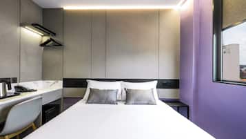 A modern hotel room with a bed, a desk with a chair, a hanging coat rack, and a window with a city view.