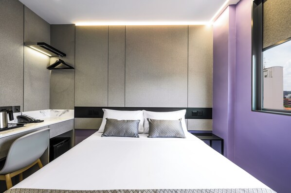 Una habitación de hotel moderna con una cama, un escritorio con silla, un perchero y una ventana con vista a la ciudad.