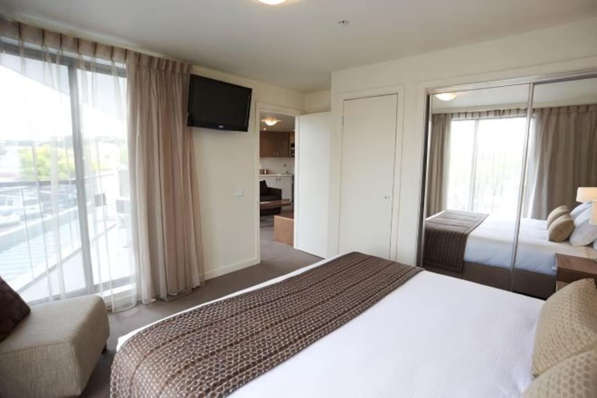 Habitación de hotel con una cama grande, televisor montado en la pared, puerta corrediza a un balcón y una pequeña zona de estar.