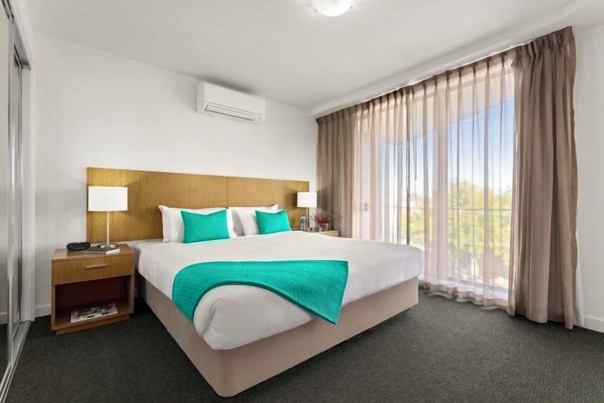 A hotel room with a large bed, two bedside tables, a nightstand, a window with curtains, and an air conditioning unit.