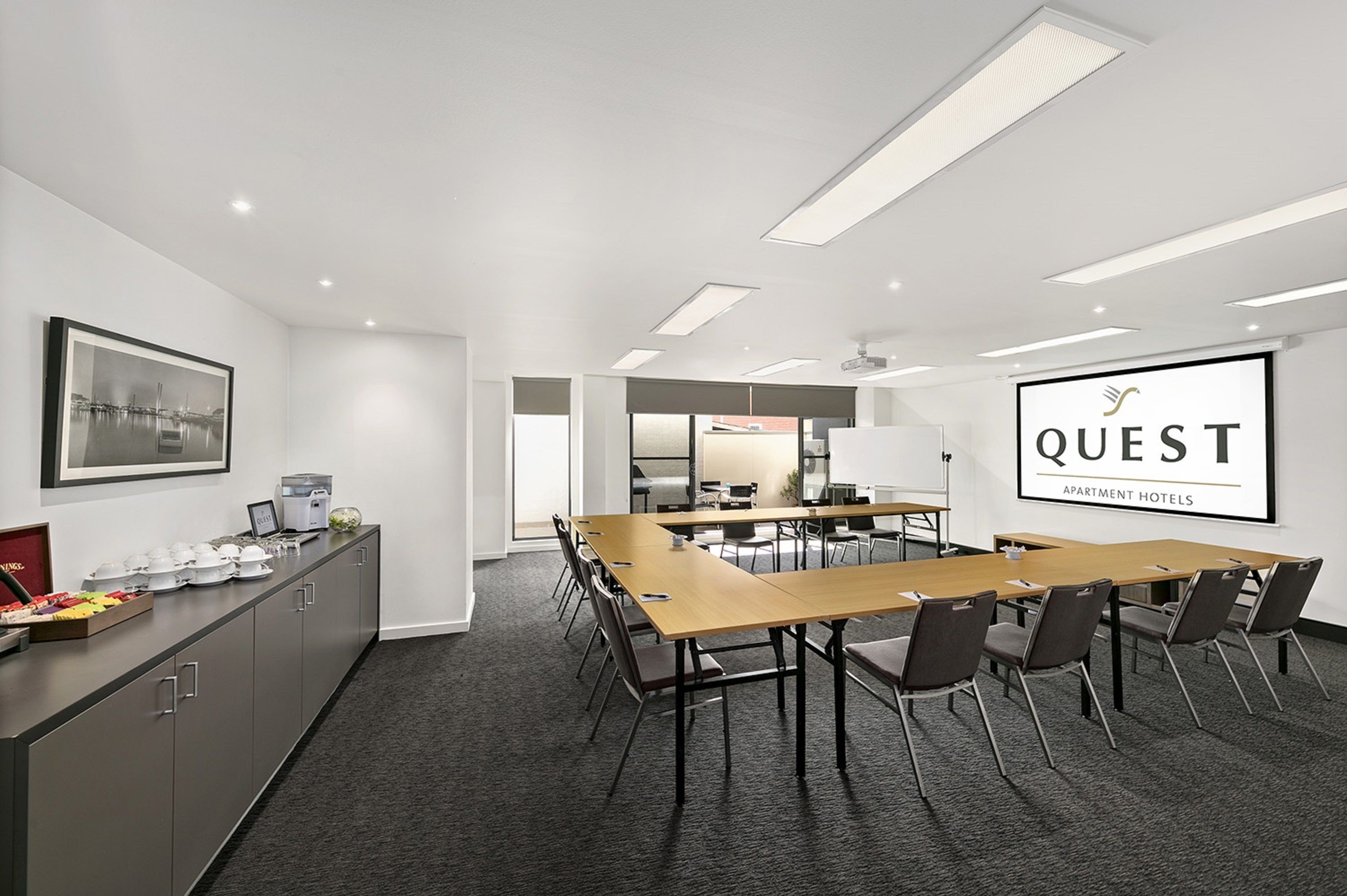 Et mødelokale med et langt bord, stole, kaffeanretning og et stort skærm, der viser "QUEST APARTMENT HOTELS".