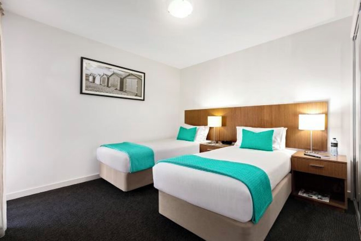 A hotel room with two beds, a wooden headboard, a nightstand, a lamp, a framed picture on the wall, and a carpeted floor.