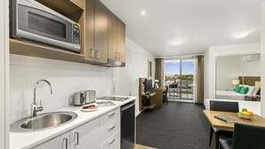 A modern kitchen with a microwave, toaster, and sink. There is a dining area with a table and chairs, and a view of the outdoors through a large window. - Quest Cheltenham (Cheltenham)