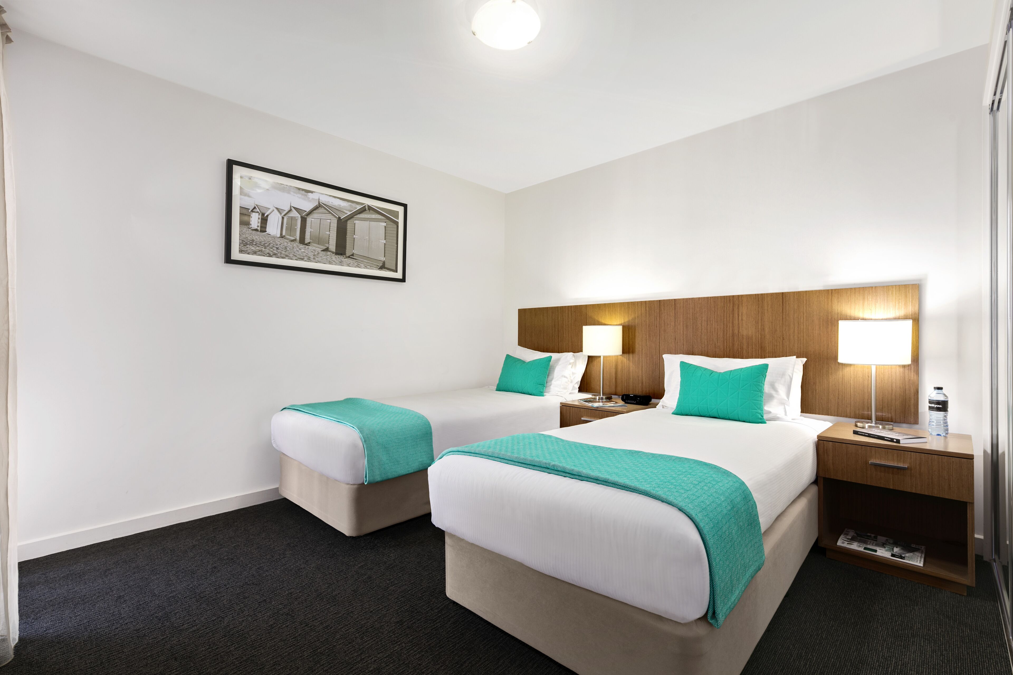 A hotel room with two beds, a wooden headboard, a nightstand with a lamp, a framed picture on the wall, and a carpeted floor.