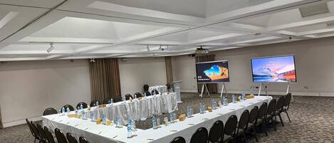 Un salón de conferencias con una mesa larga, sillas, botellas de agua y una pantalla de proyector mostrando una imagen panorámica.