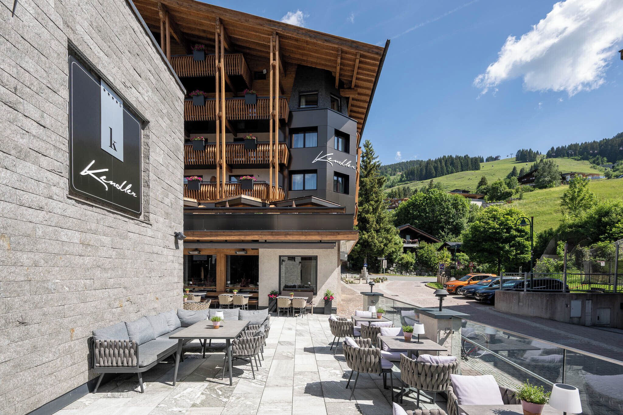 A hotel with outdoor seating, a wooden balcony, and a view of a green hillside.