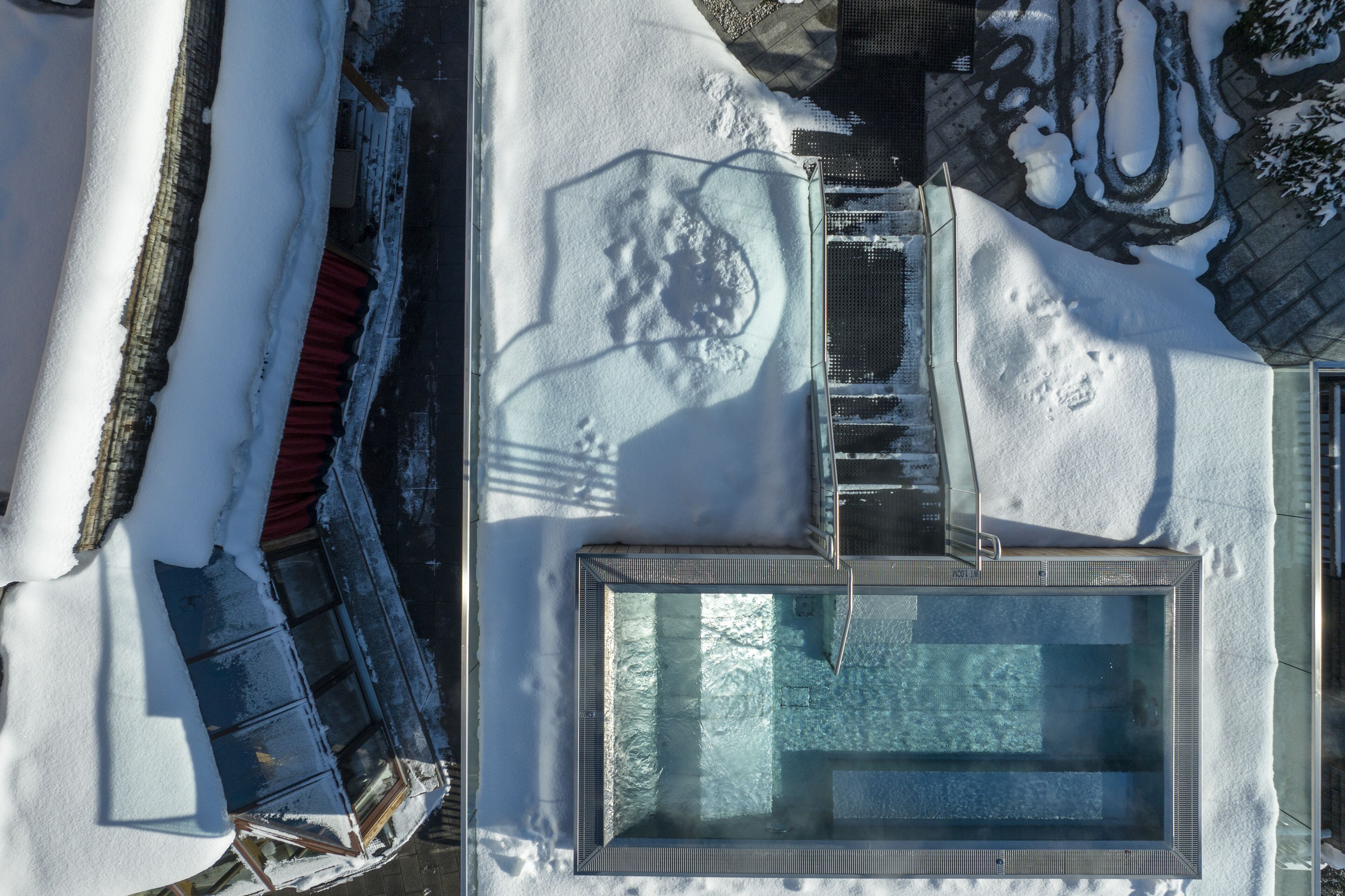A rooftop pool surrounded by snow, with a glass enclosure and a metal railing.