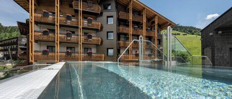 A hotel with a pool, wooden balconies with flower boxes, and a view of the surrounding landscape.