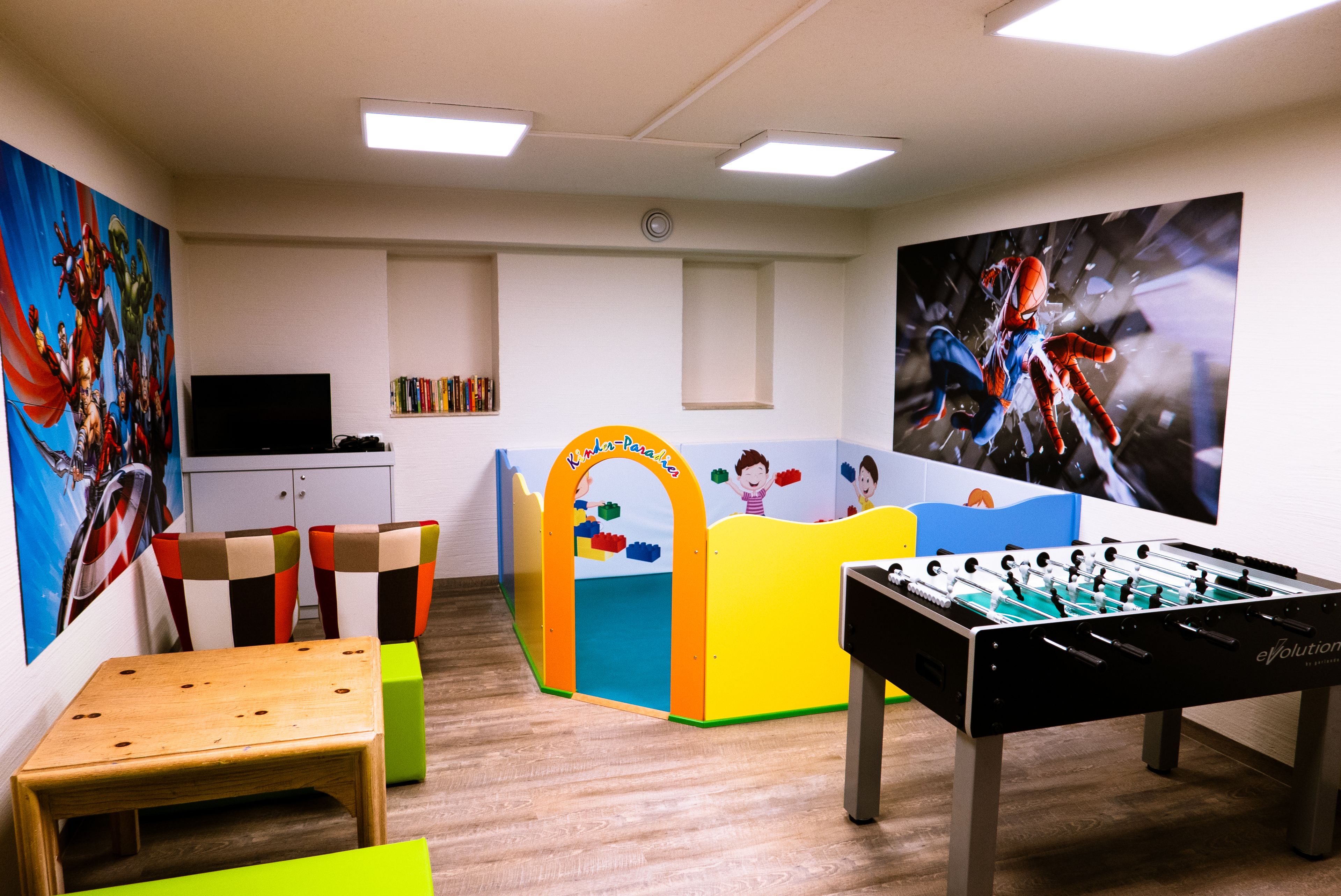 Een kamer met een tafelvoetbaltafel, een klein tafeltje, kleurrijke stoelen, een boekenkast en een televisie.