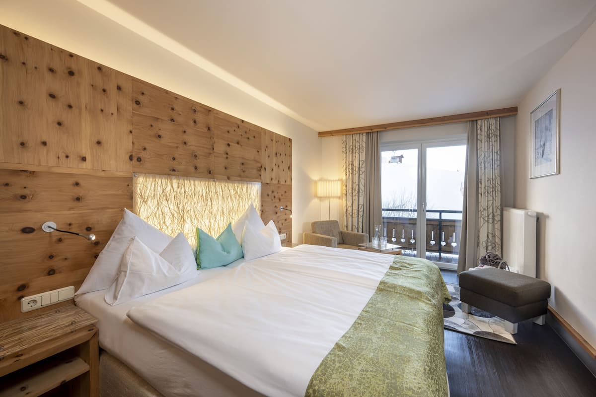 A hotel room with a large bed, a wooden headboard, a small sofa, a balcony with a view, and a window with curtains.