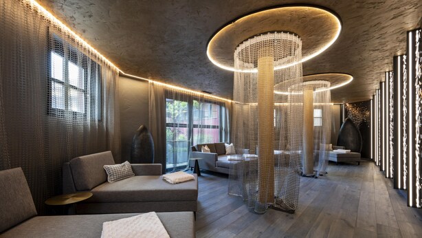 A modern interior with a wooden floor, grey sofas, and a large, circular light fixture. - Hotel Kendler (Saalbach-Hinterglemm)