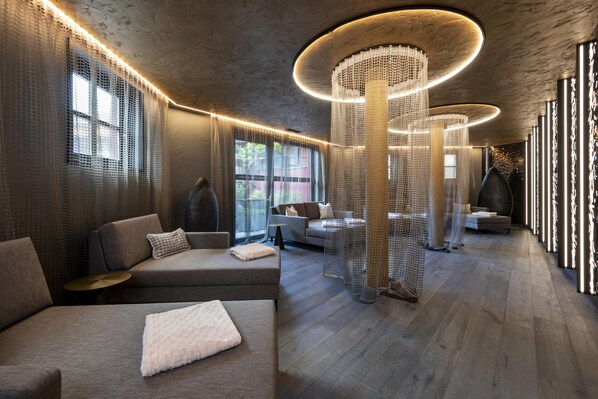 A modern interior with a wooden floor, grey sofas, and a large, circular light fixture. - Hotel Kendler (Saalbach-Hinterglemm)