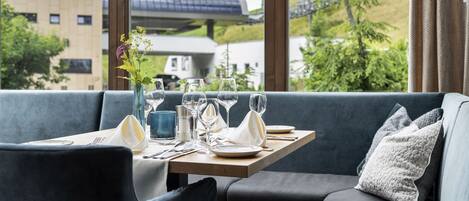 A dining area with a table set for two, a vase with flowers, and a scenic view through large windows.