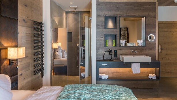A modern hotel room with a wooden ceiling, a large bed with a patterned bedspread, a glass-enclosed shower, a dark wooden vanity with a large mirror, and a bedside table with a lamp. - Hotel Kendler (Saalbach-Hinterglemm)