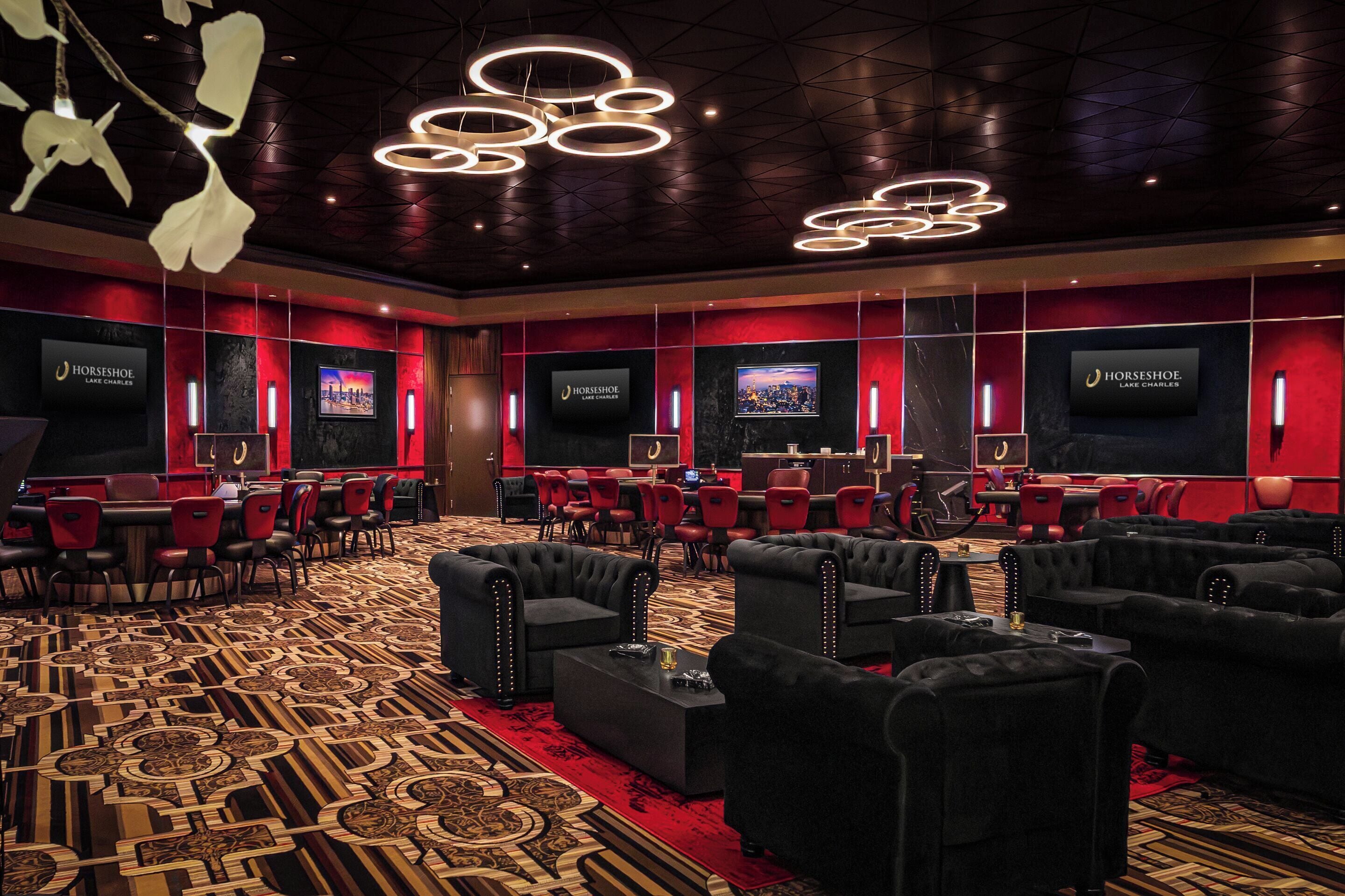 A hotel lobby with a patterned carpet, black sofas, red chairs, and multiple screens on the walls.