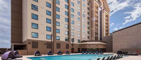 Zona de piscina de hotel con sillones de descanso, una piscina y un edificio grande con varios pisos y balcones.