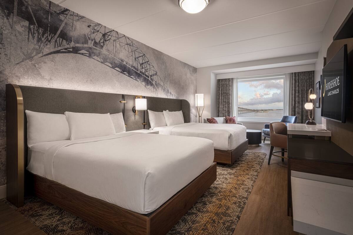 A hotel room with two beds, a desk, a chair, a TV, and a large window with a view of a bridge and water.