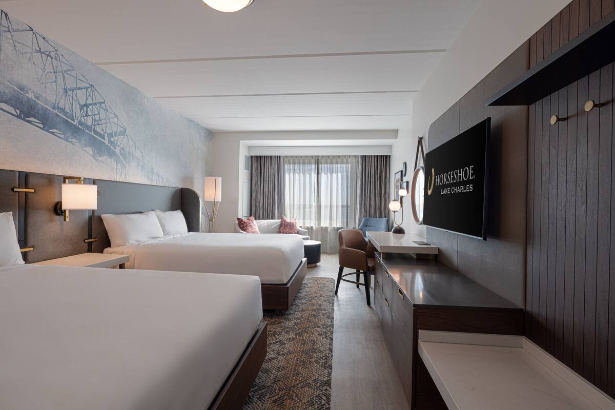 A hotel room with two beds, a desk, a chair, a TV, and a large painting on the wall.