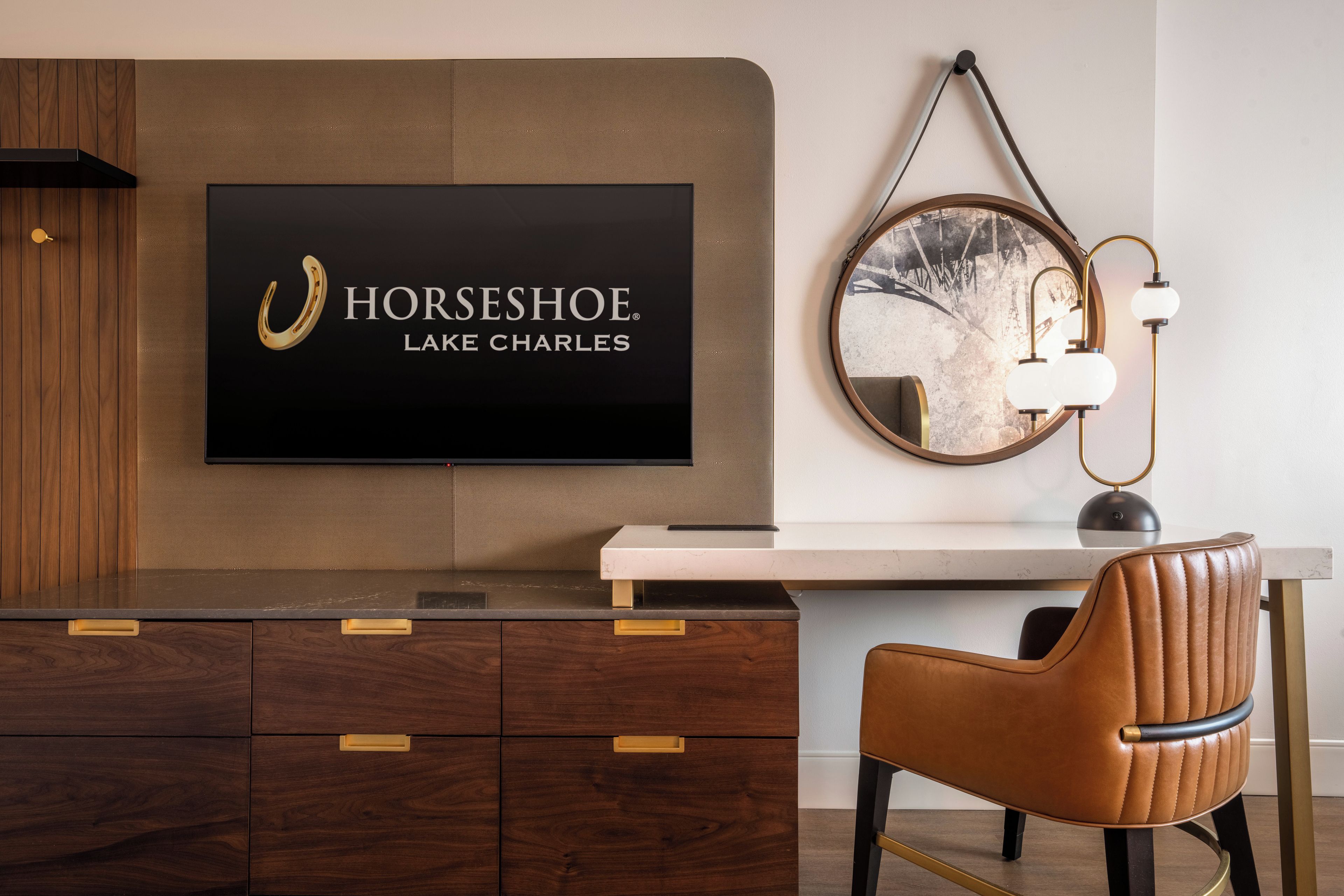 Una habitación de hotel moderna con un televisor montado que muestra el logo de Horseshoe, un espejo redondo con marco vintage, un escritorio con lámpara y una silla de cuero.