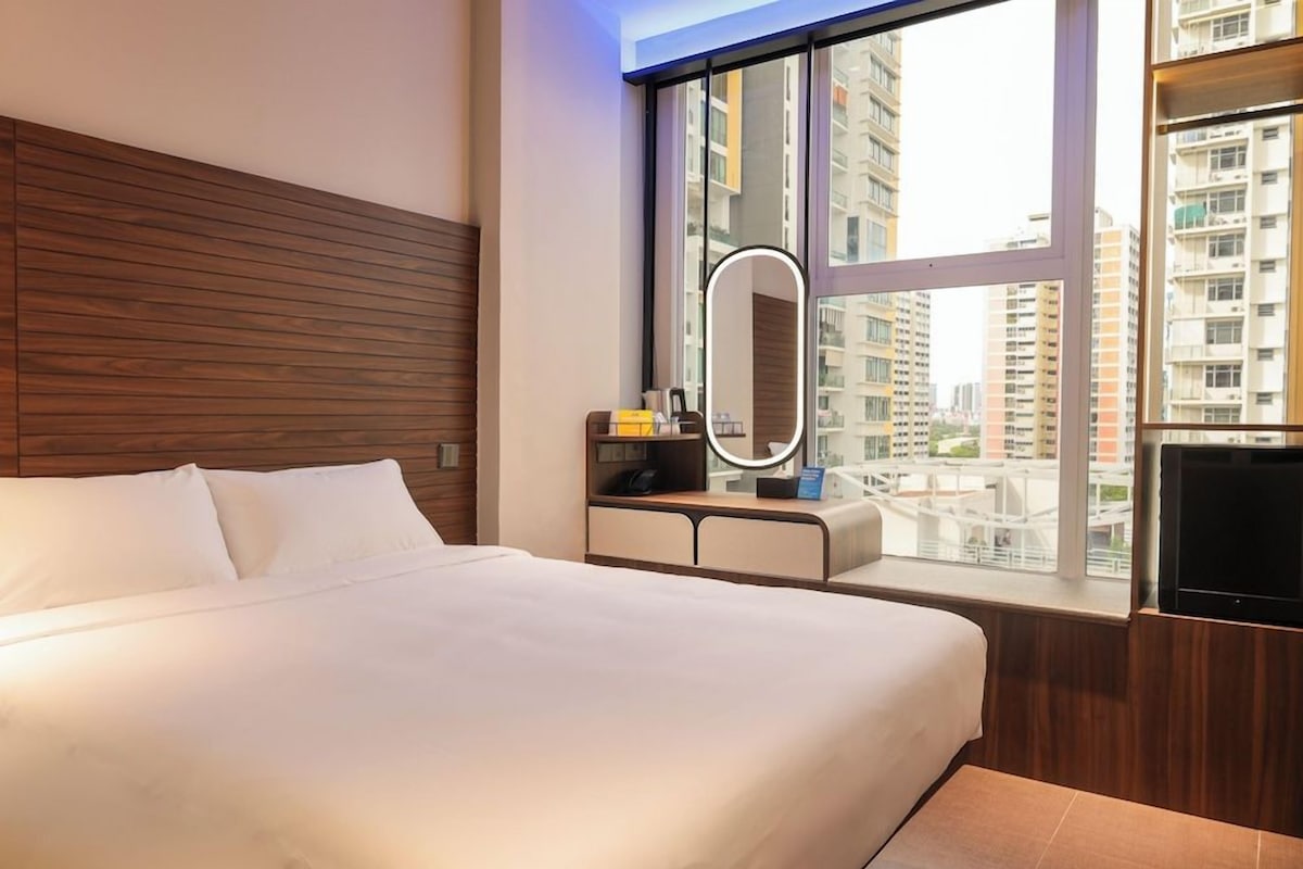 Habitación de hotel con una cama grande, una mesita de noche con espejo, un televisor y vistas a edificios altos.