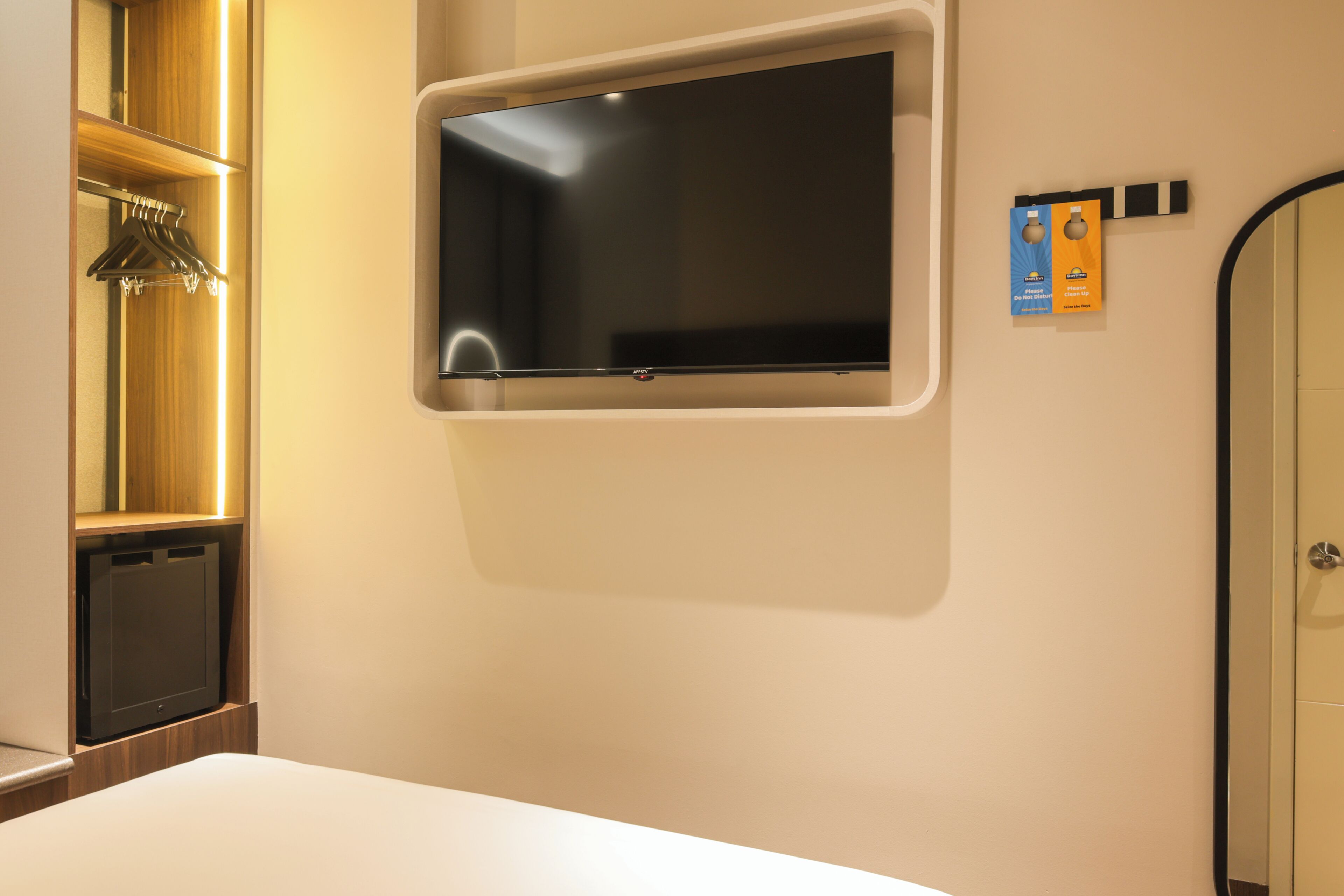 A hotel room with a mounted flat-screen TV, a wooden shelf with hanging items, and a door with a mirror.