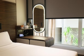 Una habitación de hotel moderna con una cama grande, una mesita de noche con espejo, un escritorio pequeño con teléfono y una ventana con vistas a zonas verdes.
