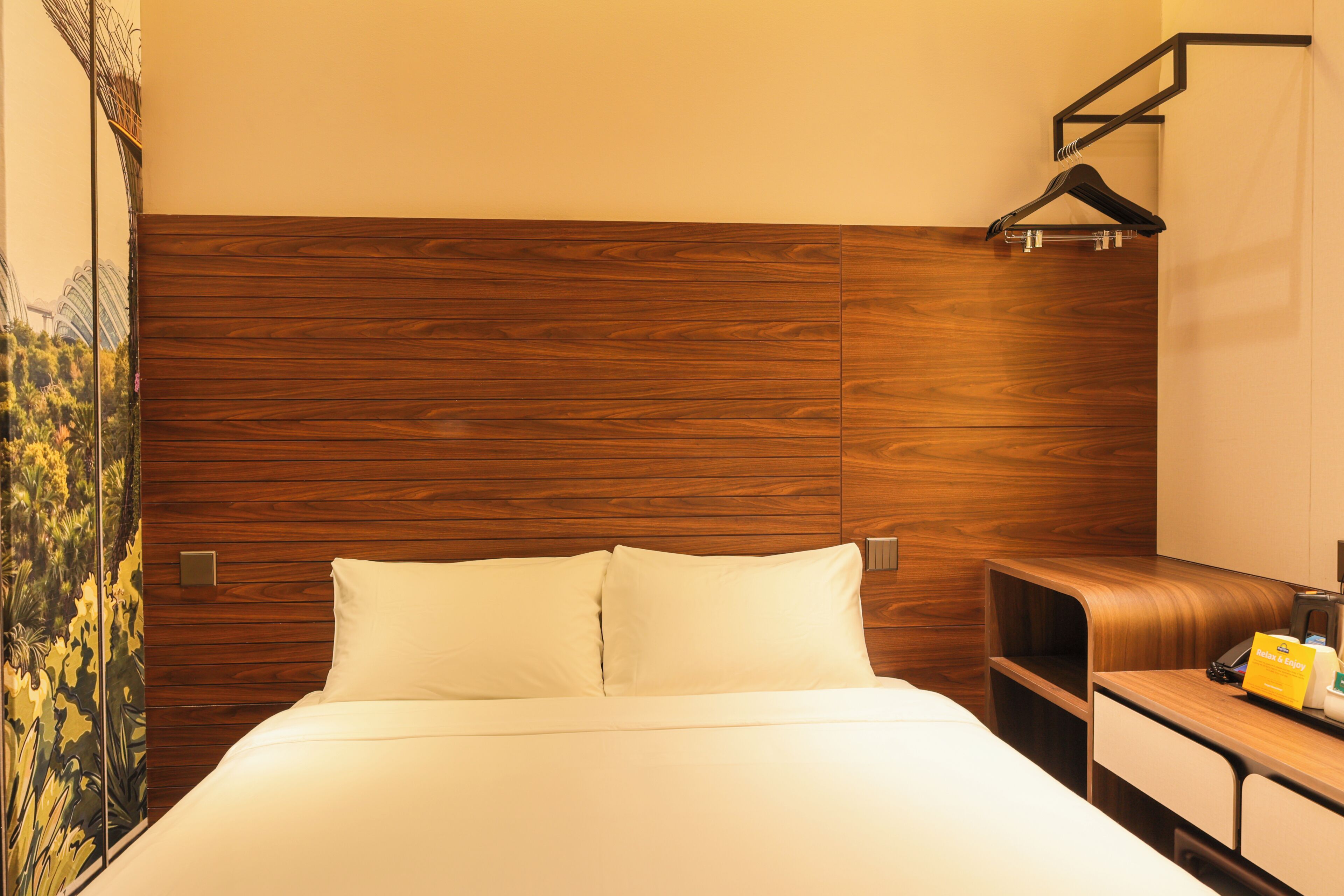 Een hotelkamer met een houten hoofdeinde, een bed met witte lakens, een nachtkastje en een klein bureau met een telefoon en een geel voorwerp erop.