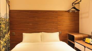 Een hotelkamer met een houten hoofdeinde, een bed met witte lakens, een nachtkastje en een klein bureau met een telefoon en een geel voorwerp erop.