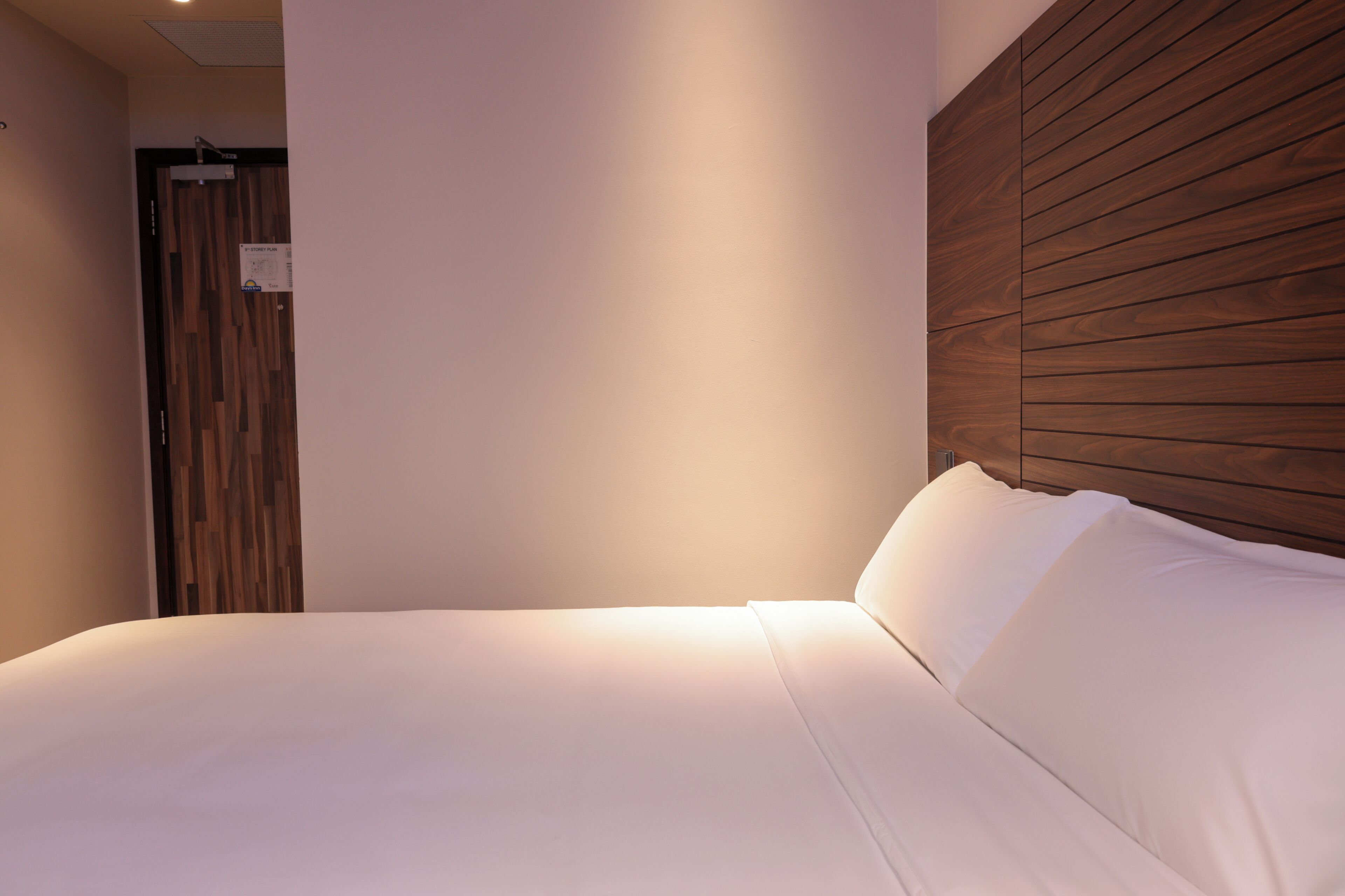 A neatly made bed with white linens, a wooden headboard, and a wall-mounted light fixture.