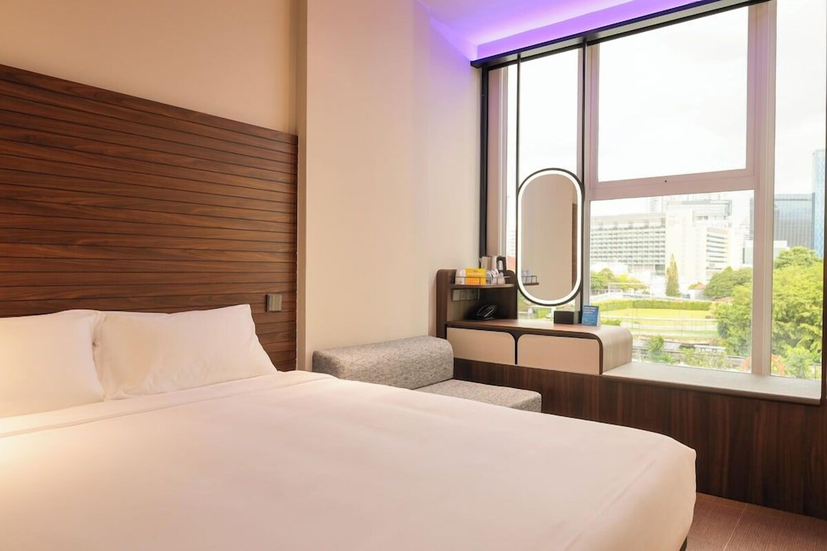A hotel room with a large bed, a small seating area, a vanity with a mirror, and a window with a city view.