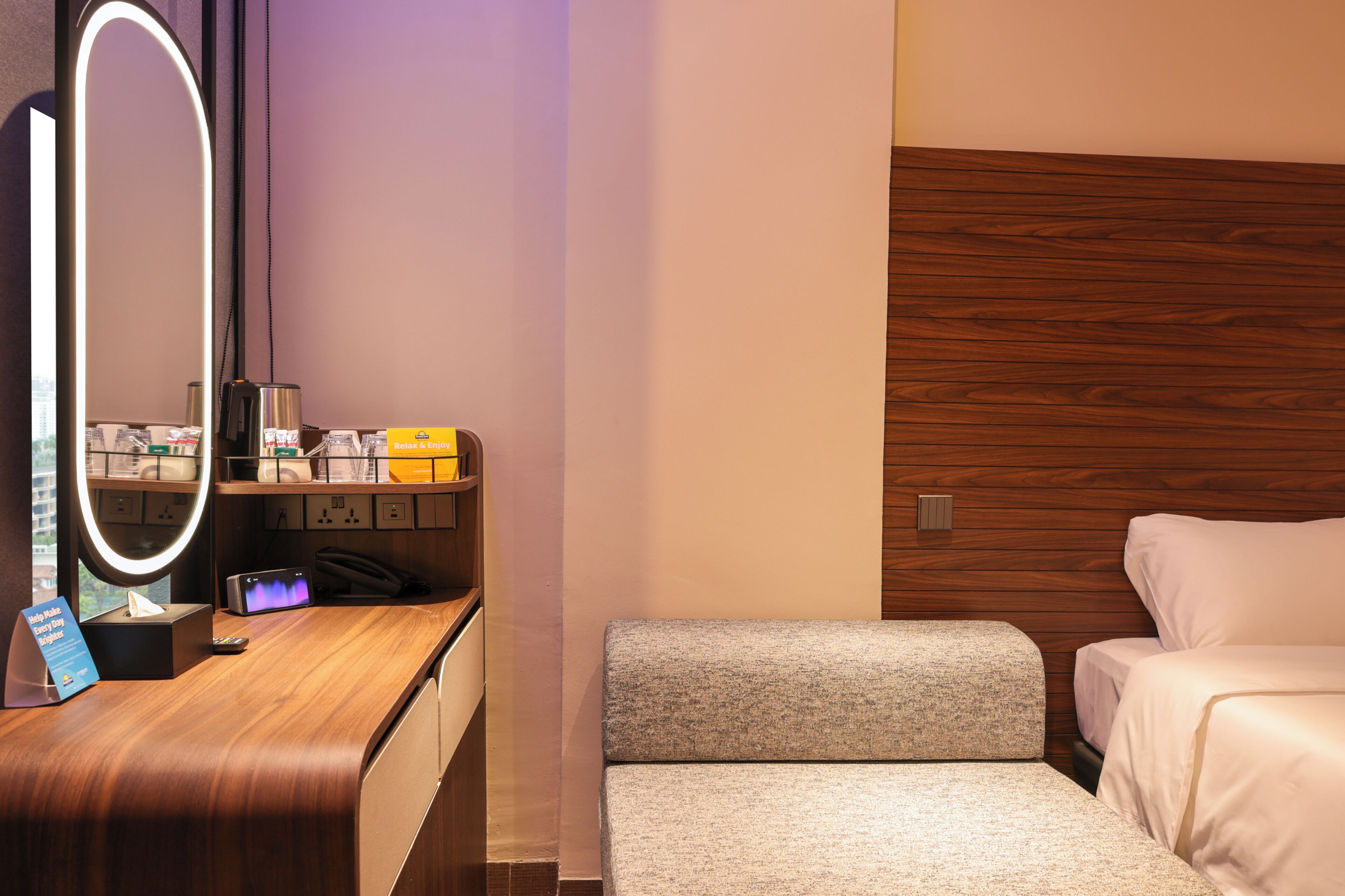 Habitación de hotel con tocador de madera, espejo, mesita de noche con artículos de aseo, una silla pequeña y cama.