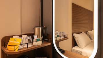 Habitación de hotel con cabecera de madera, mesita de noche con cafetera, bandeja con detalles de baño y un espejo que refleja la habitación.