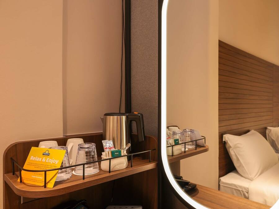 Habitación de hotel con cabecera de madera, mesita de noche con cafetera, bandeja con detalles de baño y un espejo que refleja la habitación.