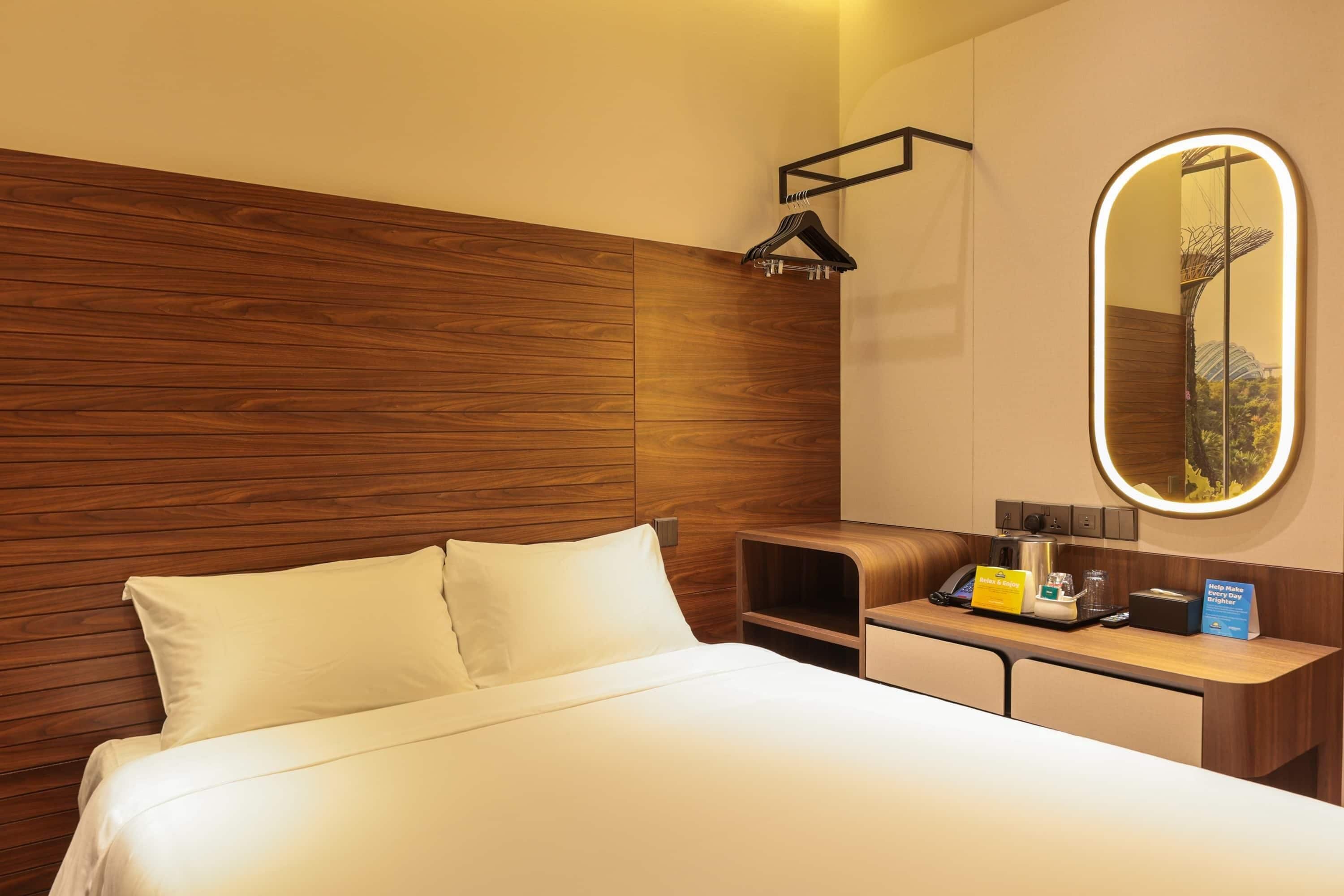 A hotel room with a wooden headboard, a bed with white linens, a bedside table with a lamp, a mirror, and a reception desk with a sign.